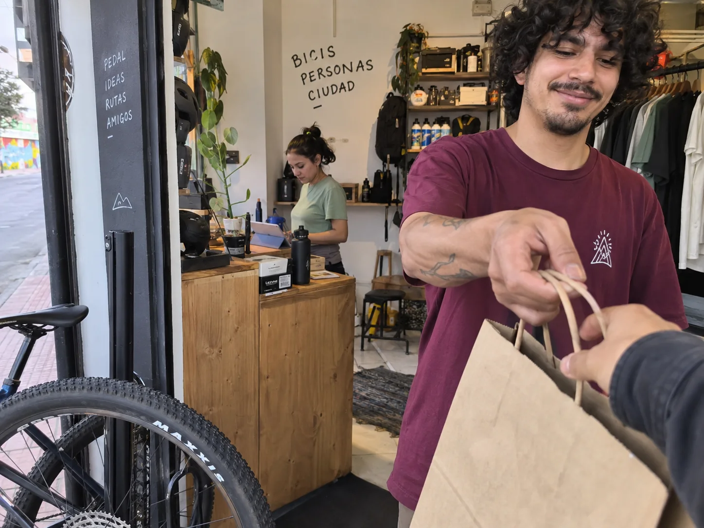 A bicycle shop owner hands a customer a paper bag, while a coworker checks an order.